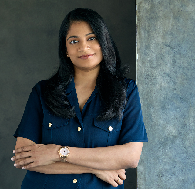 BoomBird Founder Sindhuja Sura, wearing a blue shirt with gold buttons against a gray background